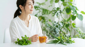 朝の光の中でハーブティーと新鮮な植物が置かれたテーブルと、リラックスした表情の女性